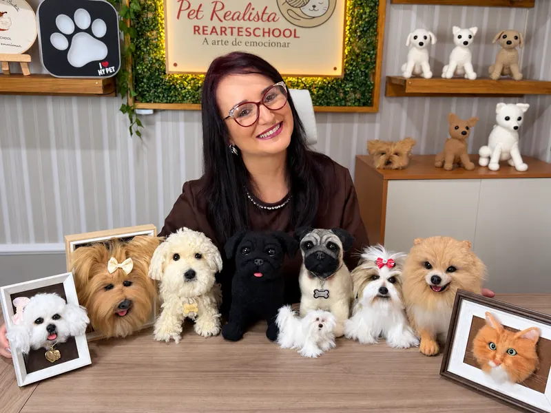 Renata Fratini à mesa do ateliê, sorrindo, atrás de nove pets realistas feitos por ela: yorkshire, poodle, dois pugs, maltês, shih-tzu, lulu e quadros com gato e bichon
